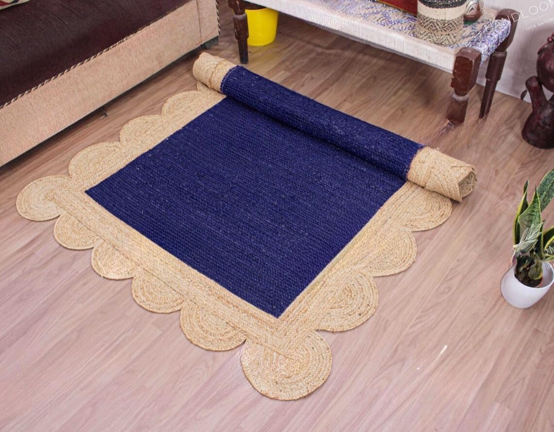 A square area rug shown partially rolled out on a light wood floor in a furnished room. The rug has a deep navy blue center with a wide natural beige braided border and scalloped edges.