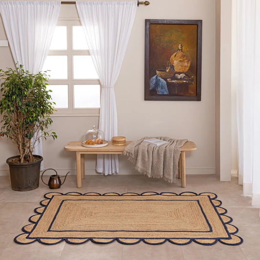Nautical-themed rug with scalloped edge on a wooden floor, with a table and plant in the background.