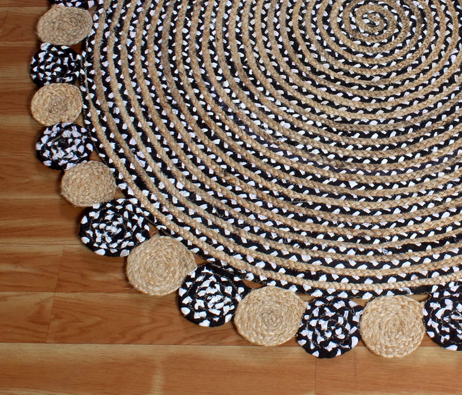 A round braided rug with a black and white patterned border, featuring alternating solid beige and black-and-white patterned tassels, laid out on a light brown wooden floor.