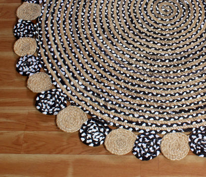 A round braided rug with a black and white patterned border, featuring alternating solid beige and black-and-white patterned tassels, laid out on a light brown wooden floor.