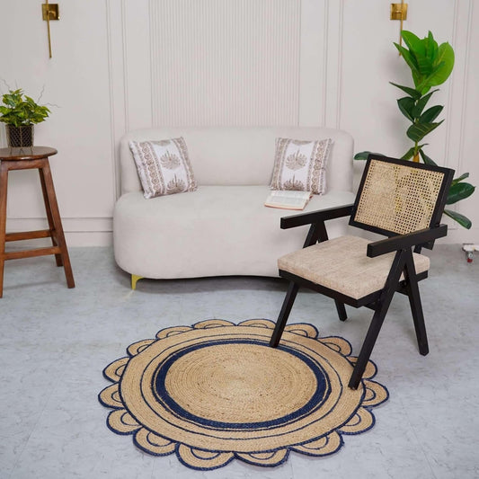 A round natural jute rug with a navy blue scalloped border, styled on a gray floor in a modern living room setting next to a cane-back wooden chair and a cream-colored sofa.