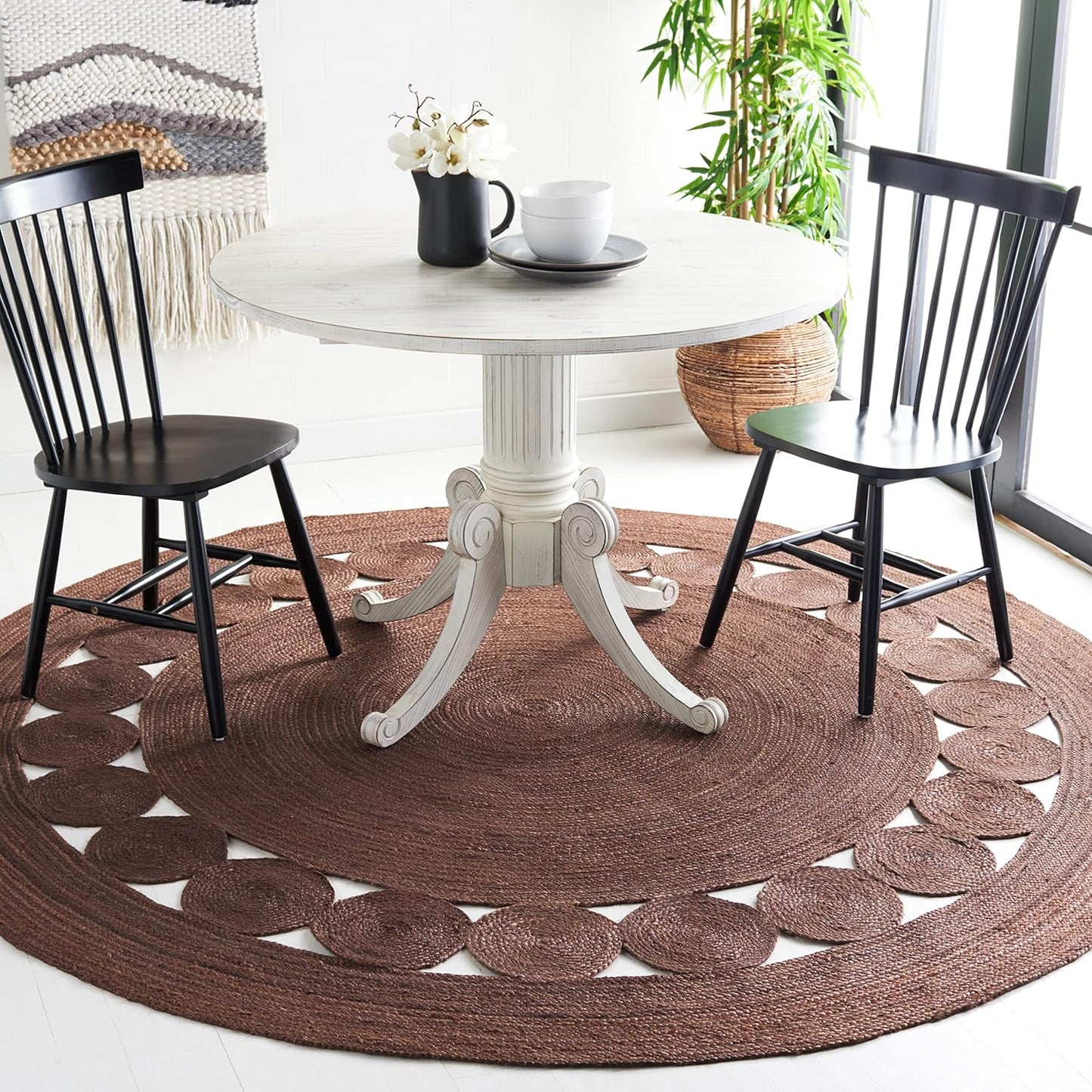 Round white dining table with intricate base, black chairs on a brown braided rug with decorative pattern.