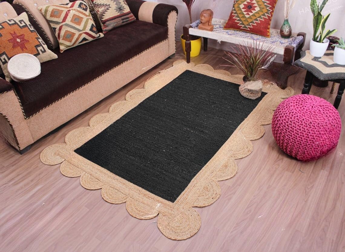 A rectangular black rug with a scalloped natural jute border, displayed on a light wood floor in a living room setting with a brown sofa, patterned pillows, a pink pouf, and a side table.