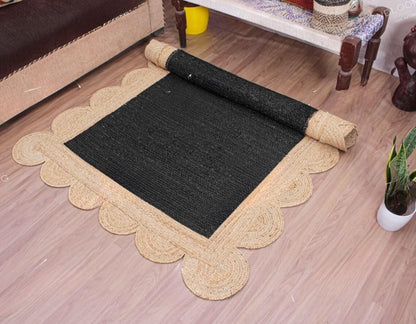 A rectangular black woven rug with a scalloped natural beige jute border, shown partially unrolled on a light wood floor in a home setting.