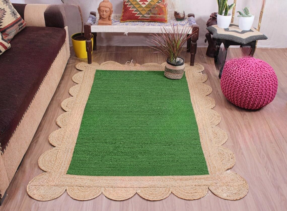 A rectangular green rug with a scalloped natural jute border, displayed on a wooden floor in a living room setting next to a brown sofa and a pink pouf.