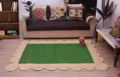 A rectangular green woven rug with a scalloped natural jute border, styled on a wooden floor in a living room setting in front of a sofa.