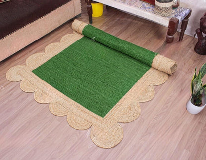 A rectangular green rug with a natural beige braided border and scalloped edges, shown partially rolled on a light wood floor in a home setting.