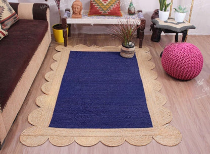A rectangular navy blue rug with a scalloped natural jute border, styled in a cozy living room setting with a brown sofa, wooden floor, and decorative accessories.