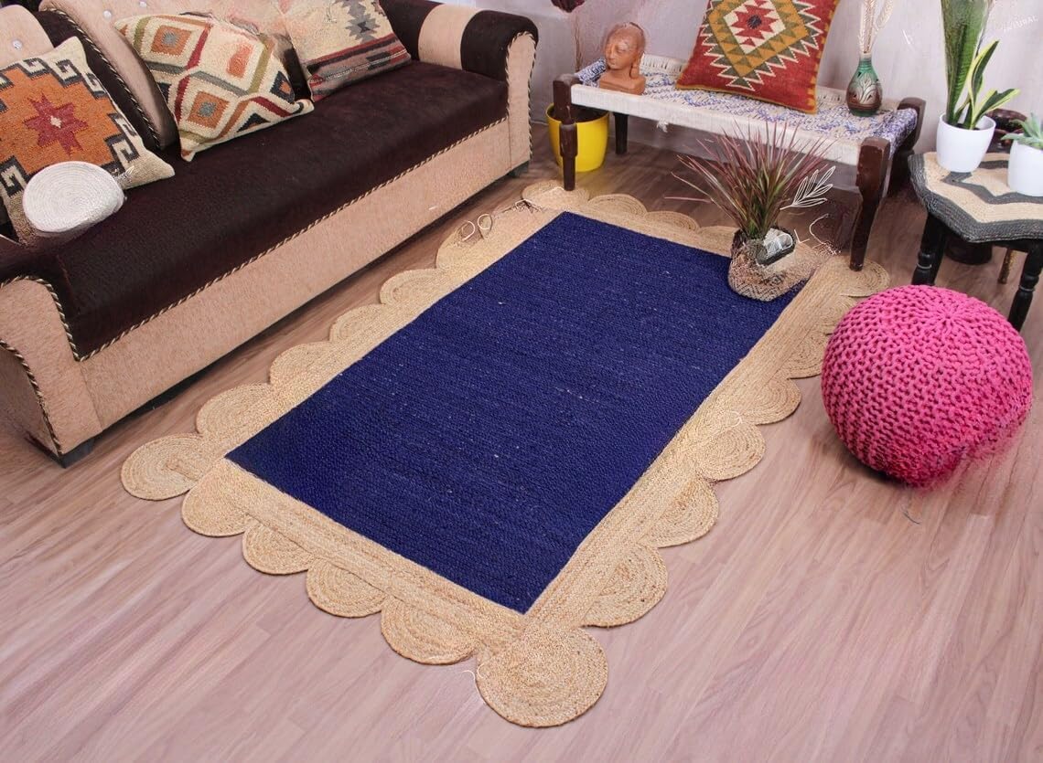 A rectangular navy blue rug with a scalloped natural jute border, styled in a living room setting on a light wood floor next to a brown sofa and a pink pouf.