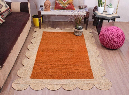 An orange rectangular area rug with a natural beige scalloped jute border, displayed on a light wood floor in a living room setting.