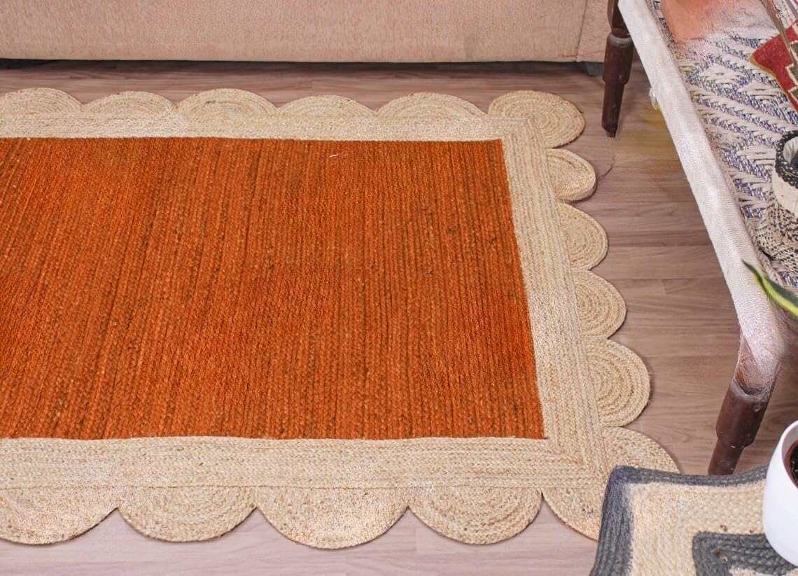 A rectangular woven rug with a warm rust-orange center and a natural beige scalloped braided border, shown on a wood floor in a living room setting.