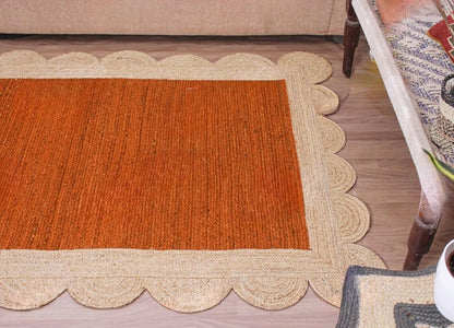 A rectangular woven rug with a warm rust-orange center and a natural beige scalloped braided border, shown on a wood floor in a living room setting.