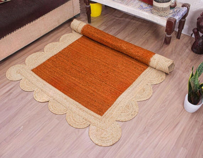 A rectangular woven rug shown partially unrolled on a light wood floor, with a rolled section revealing the backing. The rug has a warm rust-orange center and a natural beige scalloped border, placed in a styled interior setting with furniture and a small plant visible.