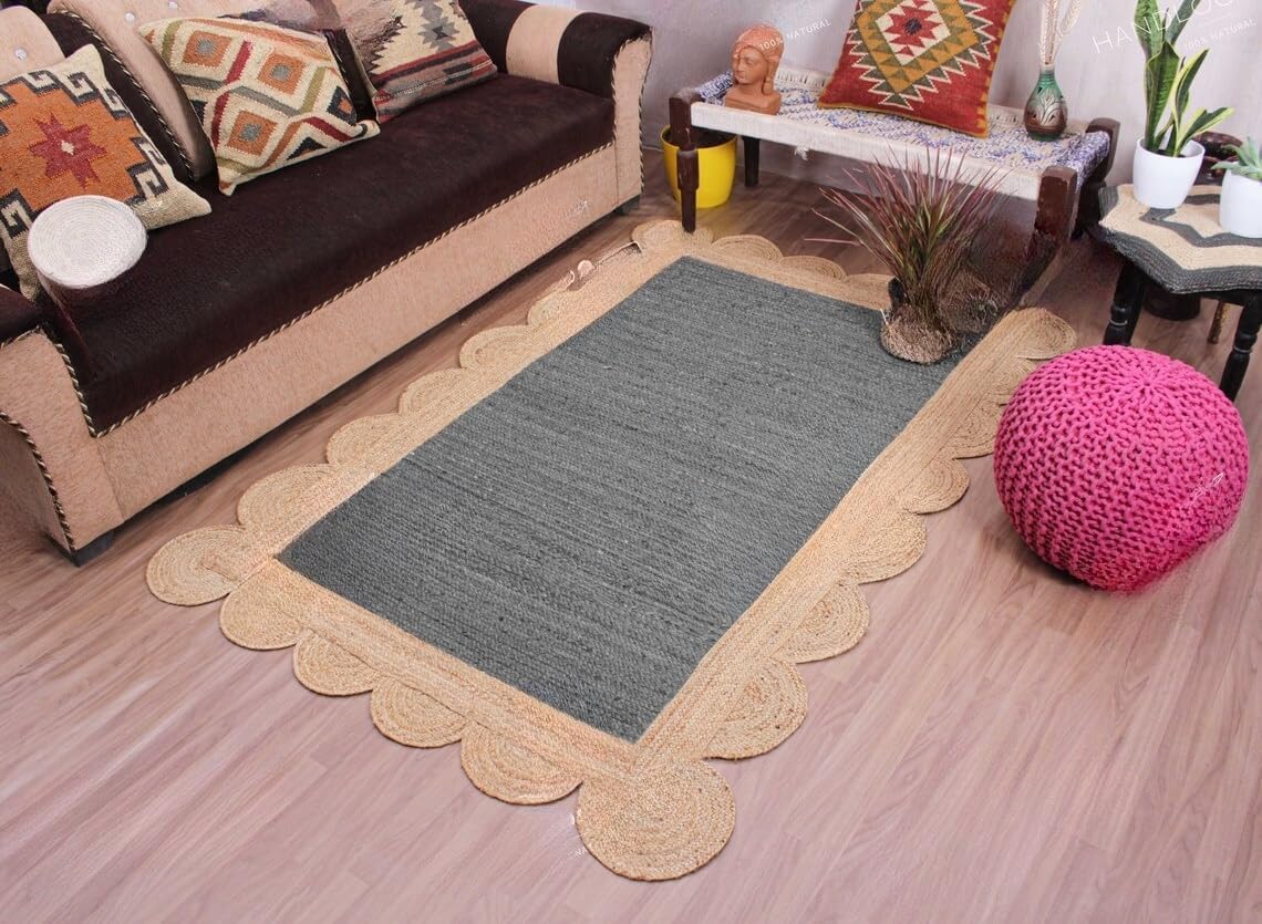 A rectangular area rug with a dark gray center and a natural beige scalloped jute border, shown in a styled living room setting with a sofa, decorative pillows, a pink pouf, and a wooden floor.