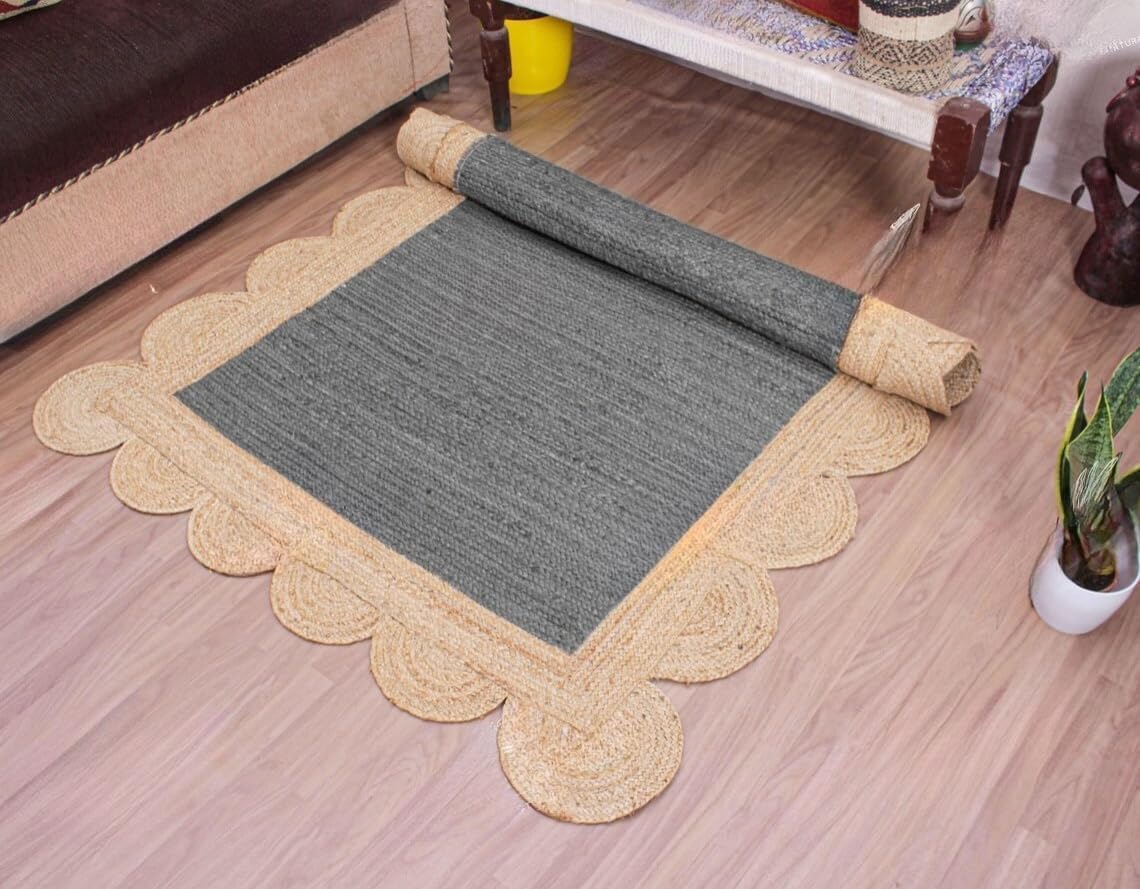 A rectangular woven rug shown partially rolled out on a light wood floor in a furnished room. The rug has a dark gray center and a natural beige scalloped border.