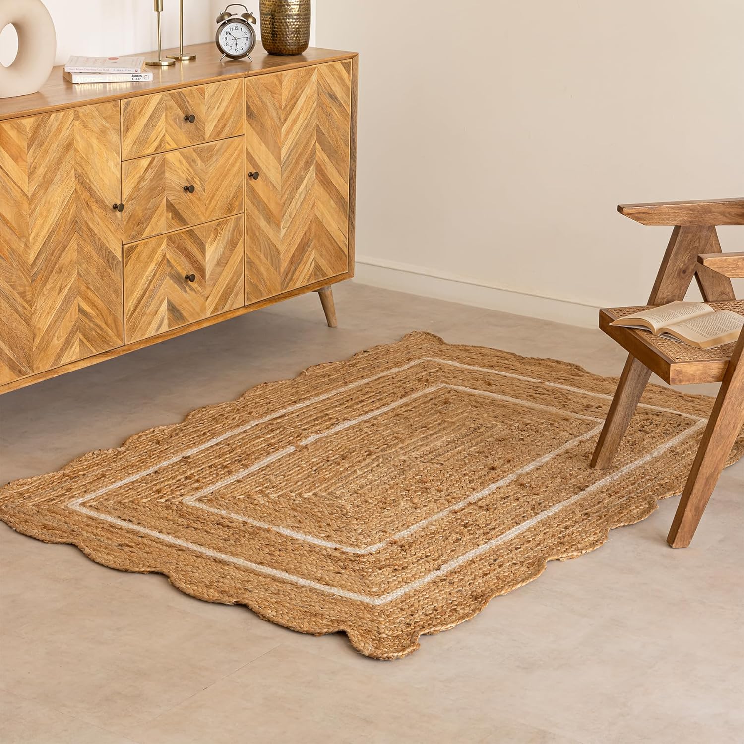 A natural jute area rug with a scalloped border and cream-colored concentric rectangular stripes, styled on a light concrete floor in a modern room next to a wooden sideboard and chair.