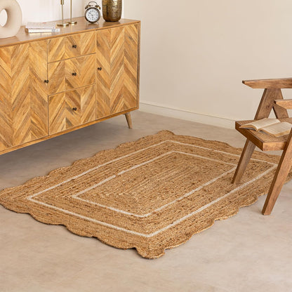 A natural jute area rug with a scalloped border and cream-colored concentric rectangular stripes, styled on a light concrete floor in a modern room next to a wooden sideboard and chair.