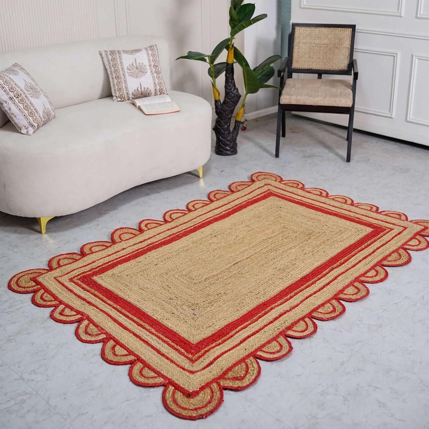 A rectangular woven rug with a natural beige center and red scalloped border, styled on a light gray floor in a living room setting next to a cream sofa and a chair.