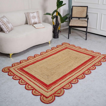 A rectangular woven rug with a natural beige center and red scalloped border, styled on a light gray floor in a living room setting next to a cream sofa and a chair.