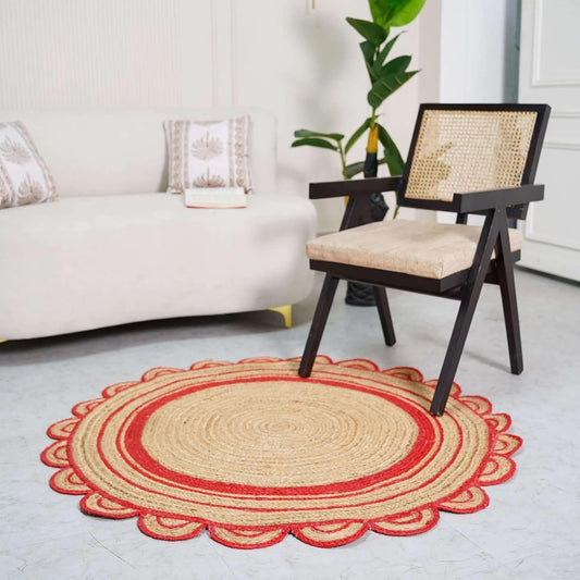 A round braided area rug with a red scalloped border and beige center, styled in a bright living room next to a wooden armchair.
