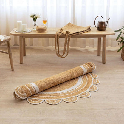 A round jute rug with a scalloped edge, shown partially unrolled on a light wood floor in a styled room setting with a wooden bench and table in the background.