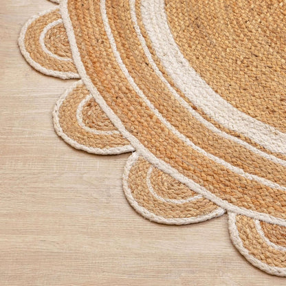 A close-up of a round braided rug with a scalloped edge, shown on a light wood floor. The rug features concentric rings in natural tan and cream.