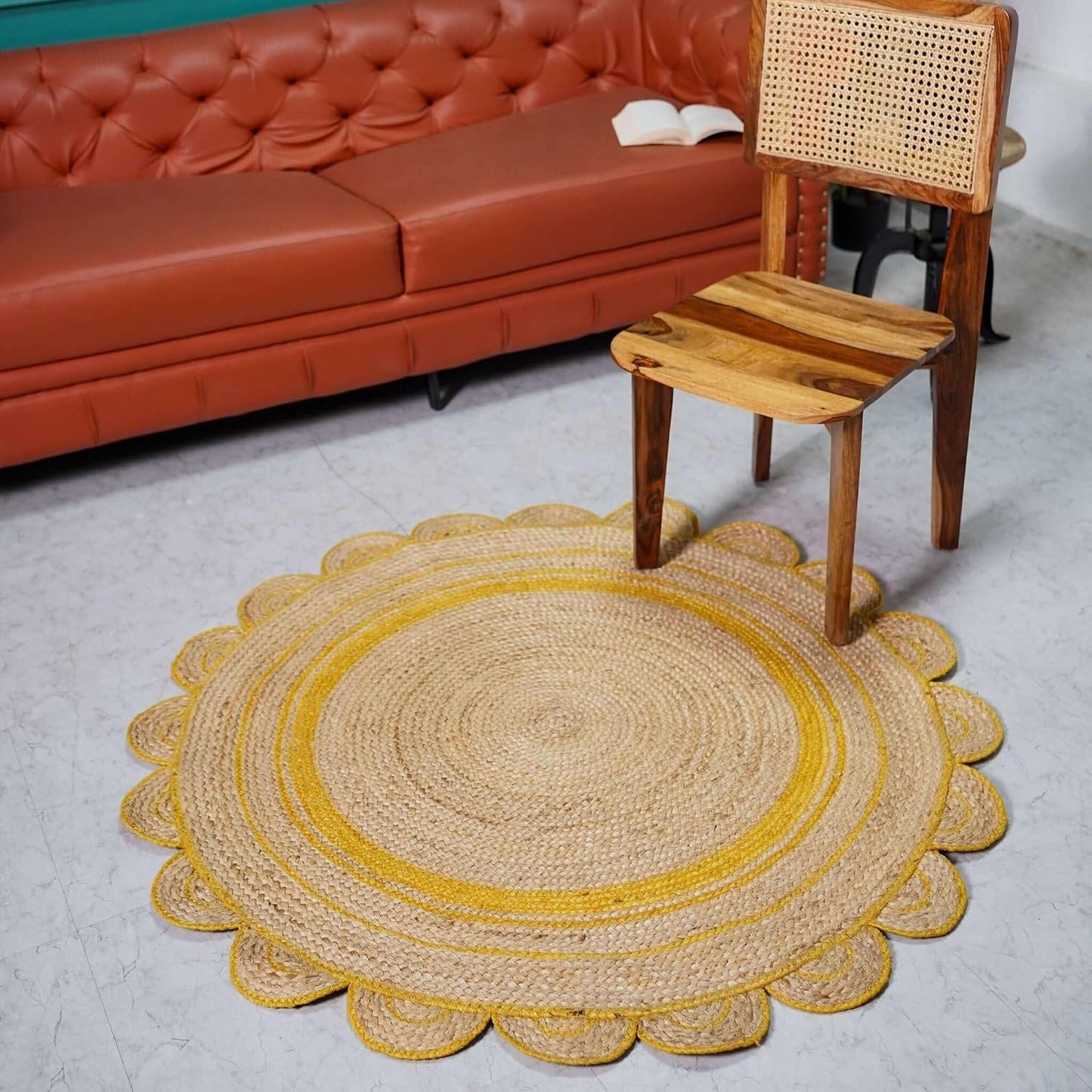 A round braided jute rug with a scalloped edge in natural beige and mustard yellow, styled on a light gray floor in a living room setting next to a rust-colored leather sofa and a wooden chair.