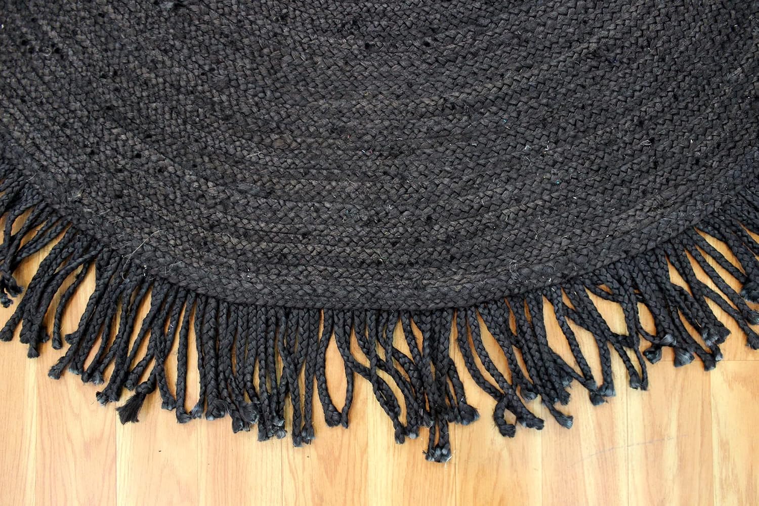 A close-up of a dark charcoal gray braided rug with a fringed edge, laid out on a light-colored wooden floor.