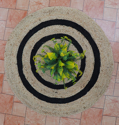 Round woven mat with black border and green plant on a tiled floor