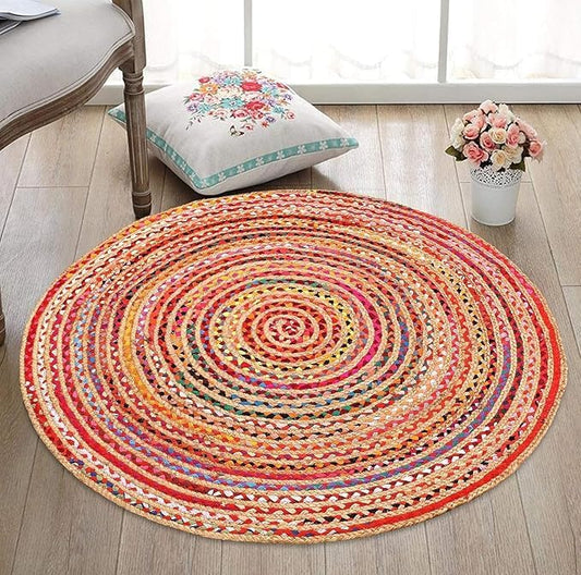 Round multicolored braided rug on a wooden floor with a pillow and flowers in the background.